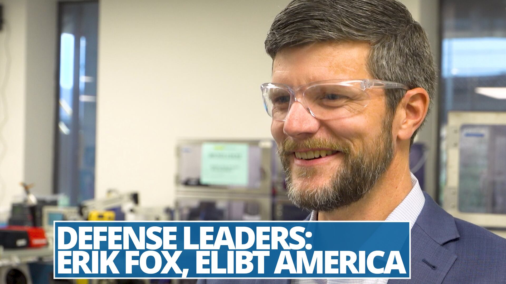 A man with short, neatly styled dark hair and a beard is smiling while wearing clear safety glasses. He is dressed in a blue suit jacket over a light-colored shirt and appears to be in a professional or industrial setting, with blurred equipment and machinery visible in the background. The environment suggests a workplace focused on technology or manufacturing.